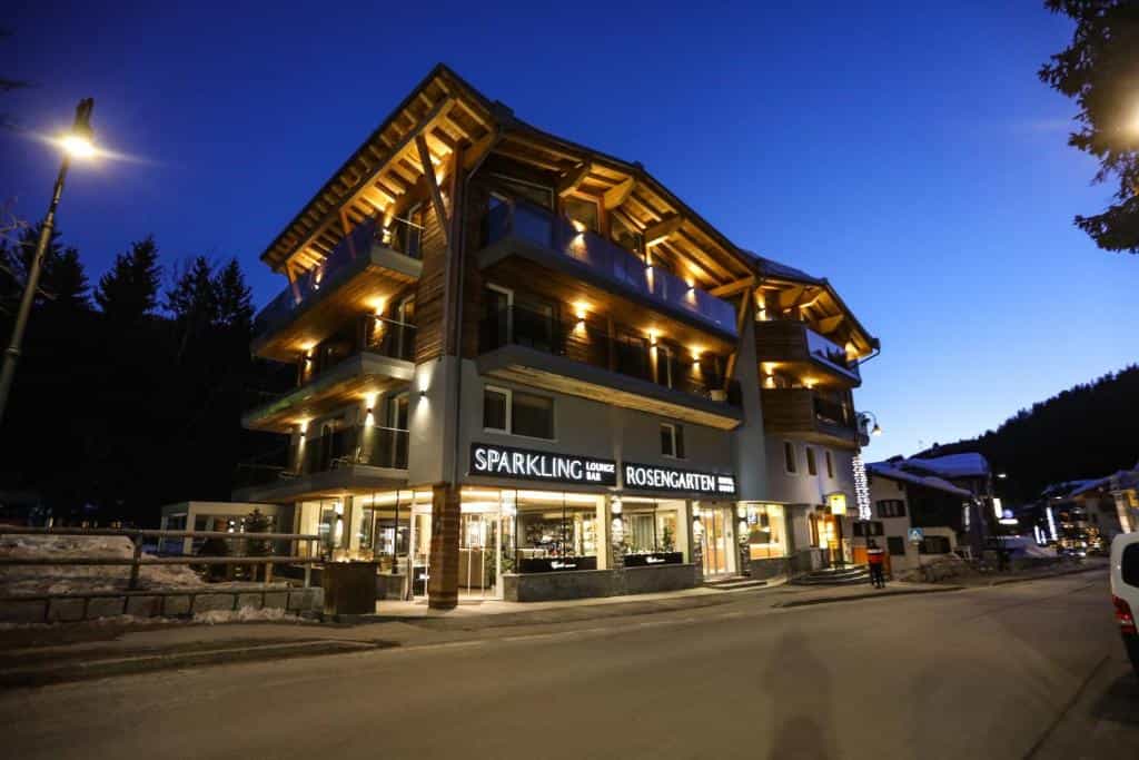 Illuminated hotel with "Sparkling Lounge Bar Rosengarten" sign at dusk in a snowy mountain village.