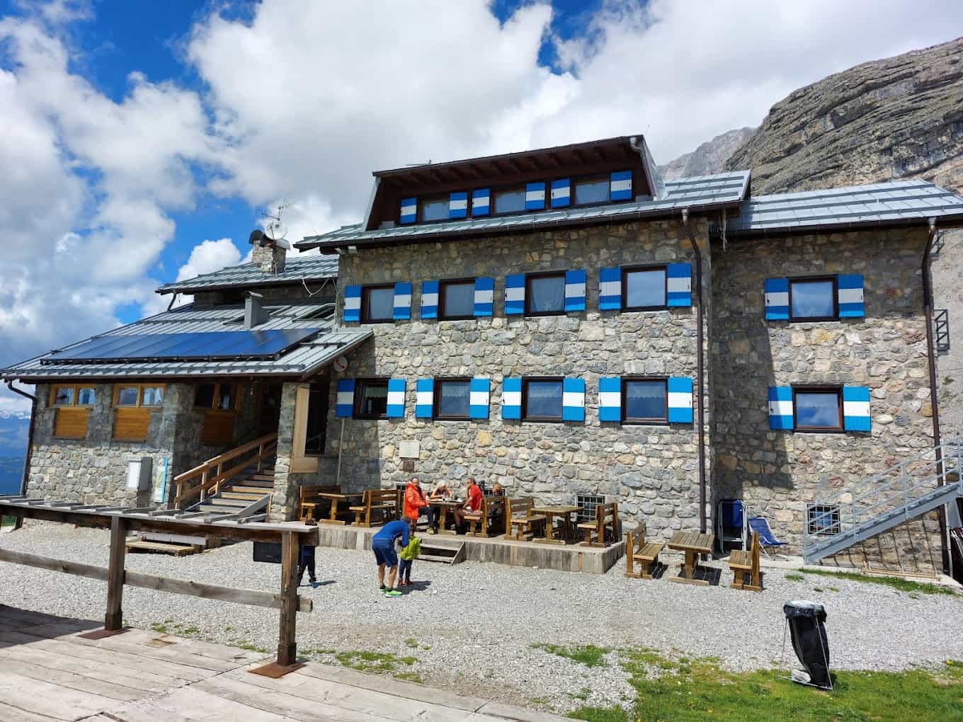 Stone mountain refuge with blue and white shutters, hikers resting outside under cloudy sky.