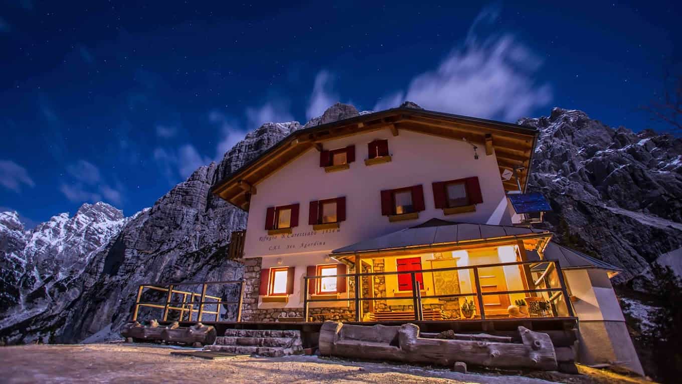 Rifugio Carestiato mountain hut illuminated at night under a starry sky with snowy peaks.