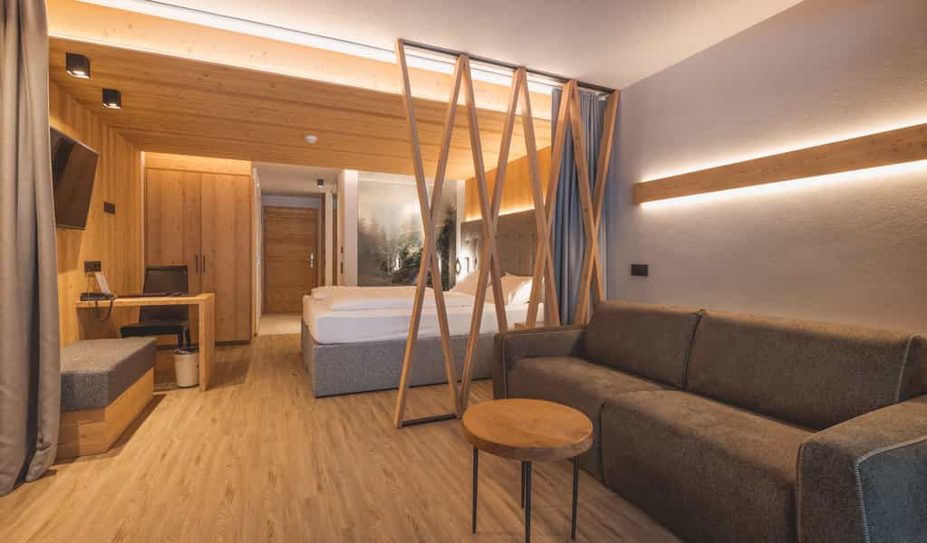 Modern hotel room with wood paneling, sofa, desk, and bed separated by wooden X-frame.