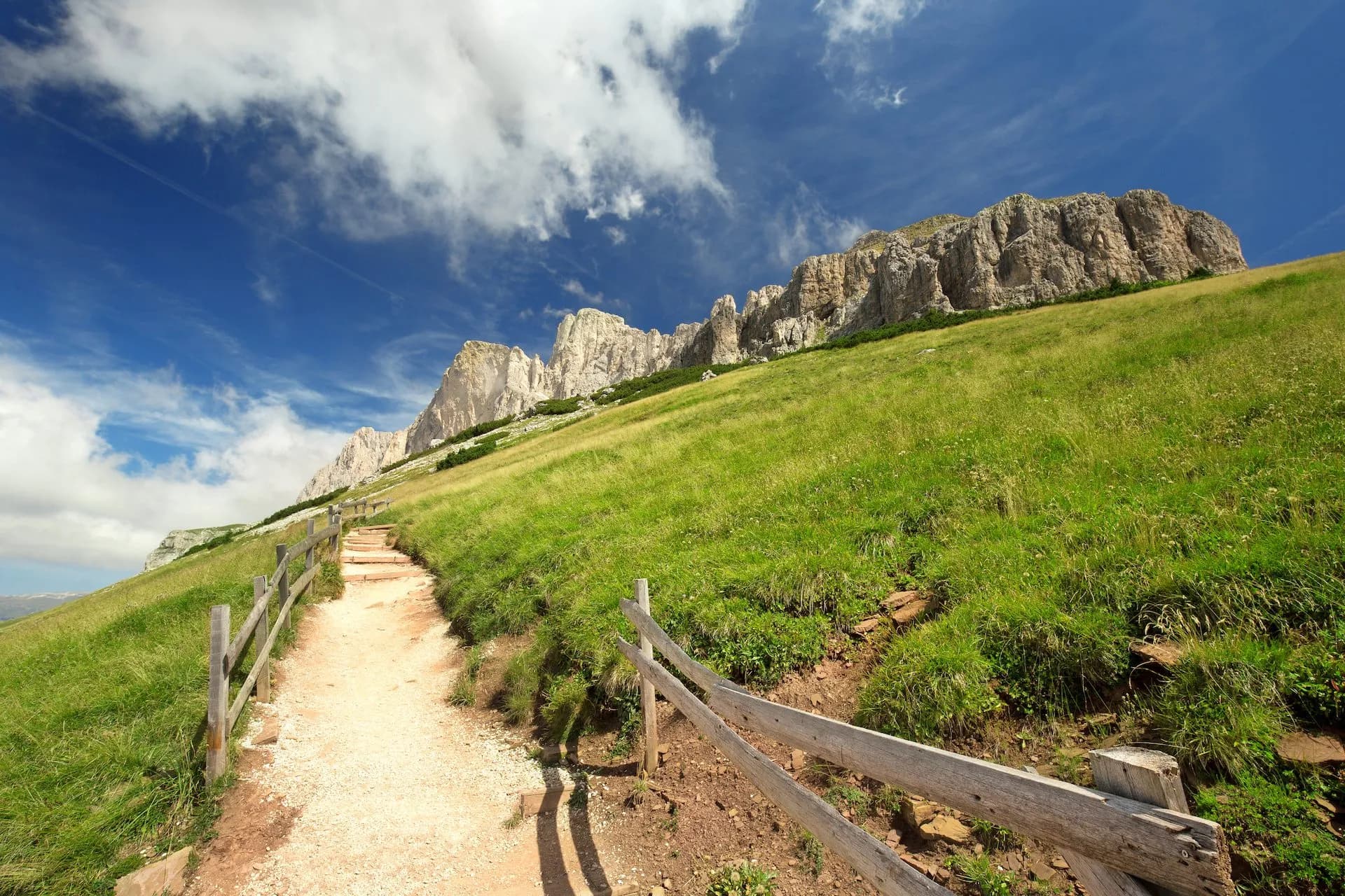 Hiking path up a grassy slope toward rocky Dolomite mountains under a blue sky