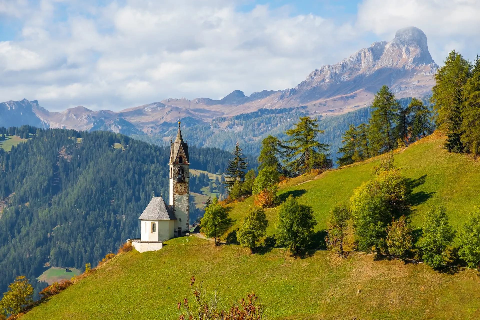 Church on grassy hillside with pine forest and rugged mountains in La Val Badia.