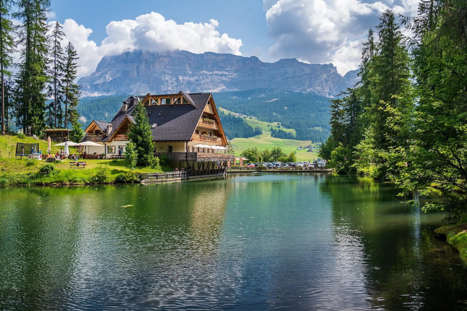 Lakeside chalet next to dark water with tall pine trees and rugged mountains under clouds.