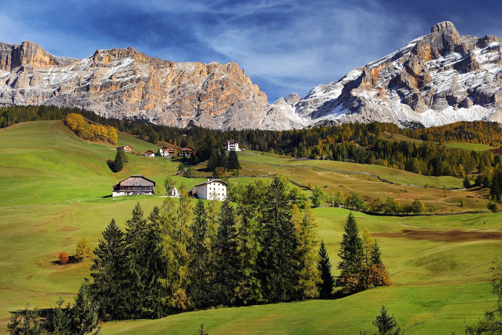 Val Badia Dolomites with green meadows, autumn trees, and snow-dusted rocky mountains under blue sky.