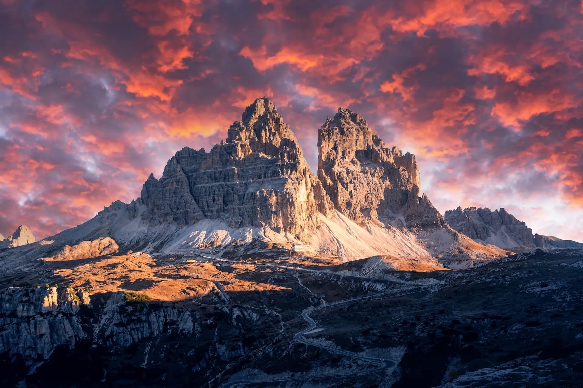 Tre Cime di Lavaredo mountains at sunset with dramatic red clouds and winding road below.