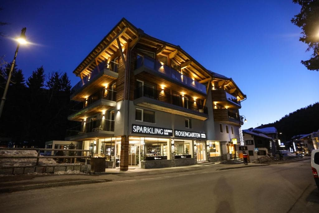Illuminated hotel with "Sparkling Lounge Bar Rosengarten" sign at dusk in a snowy mountain village.