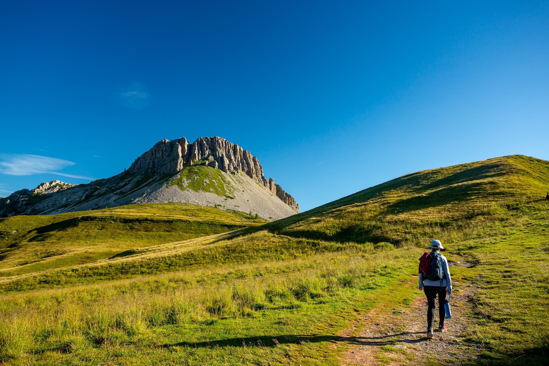 Hiker walking on dirt path toward steep rocky mountain under clear blue sky, Monte Castellaz Loop.