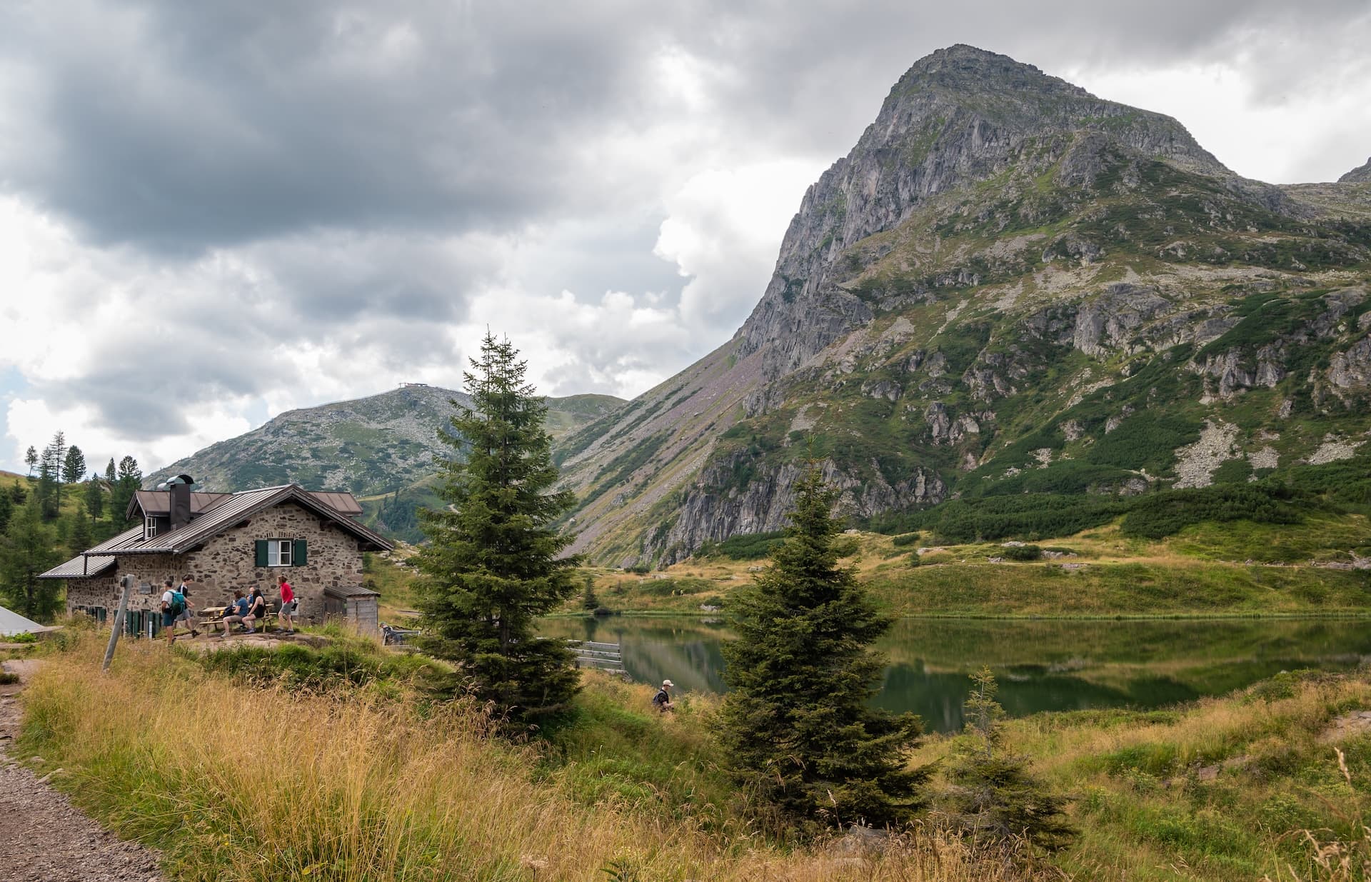Rifugio at Colbricon Lakes with hikers resting by stone hut and steep mountain.