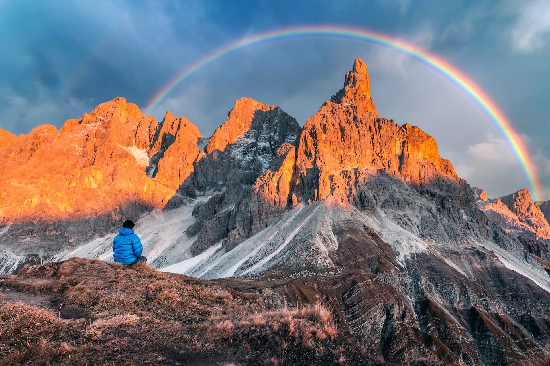 Hiker watching sunset light on Pale di San Martino mountains under a rainbow.