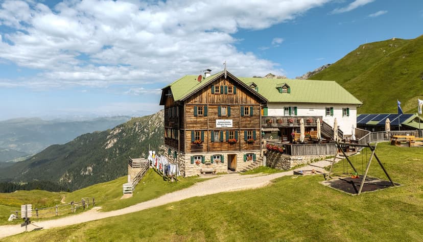Alpine mountain hut Rifugio Genova with green roof on grassy slope overlooking distant peaks.