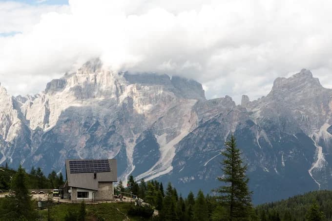 Mountain cabin with solar panels nestled in front of rugged alpine peaks under a cloudy sky.