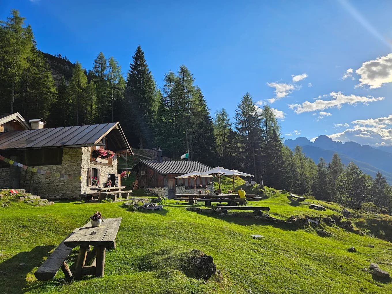 Stone mountain huts with outdoor dining tables on a grassy alpine meadow surrounded by pine forests and mountain peaks.