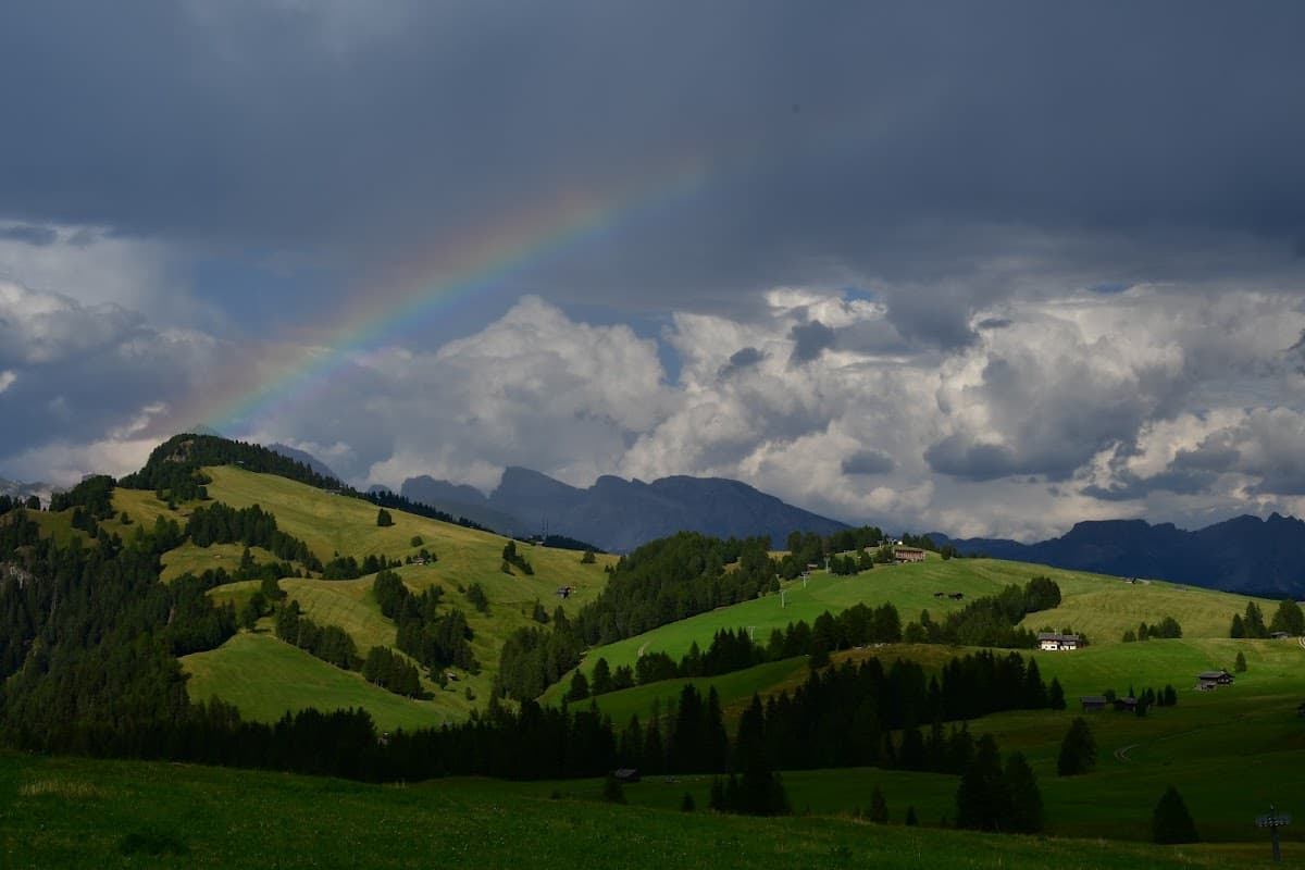 Rainbow over green alpine hills with scattered trees and mountains under a cloudy sky