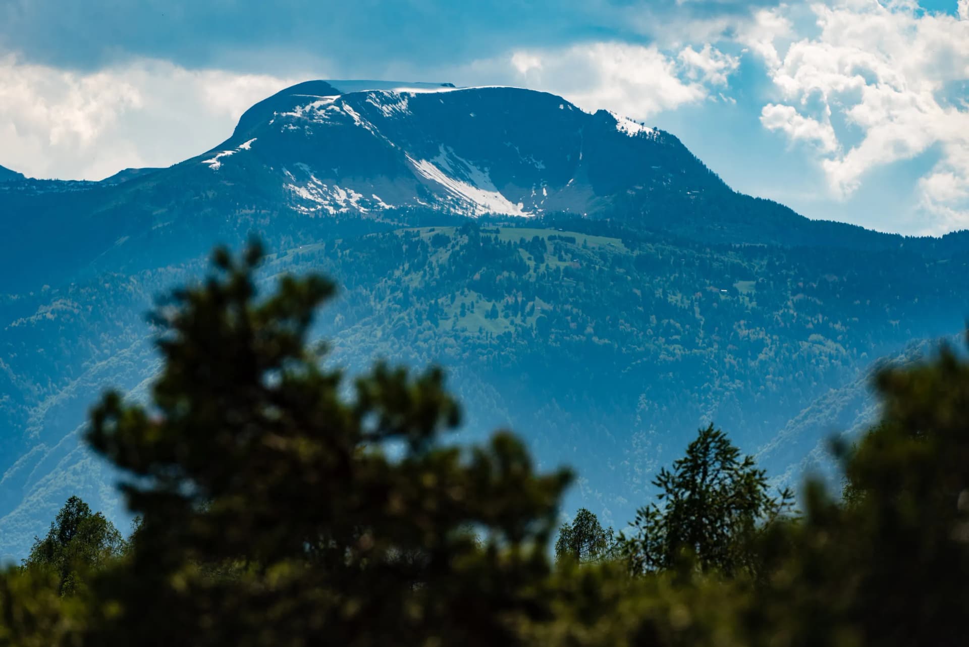 Mountain peak with residual snow cap viewed over hazy, forested foothills under blue sky.