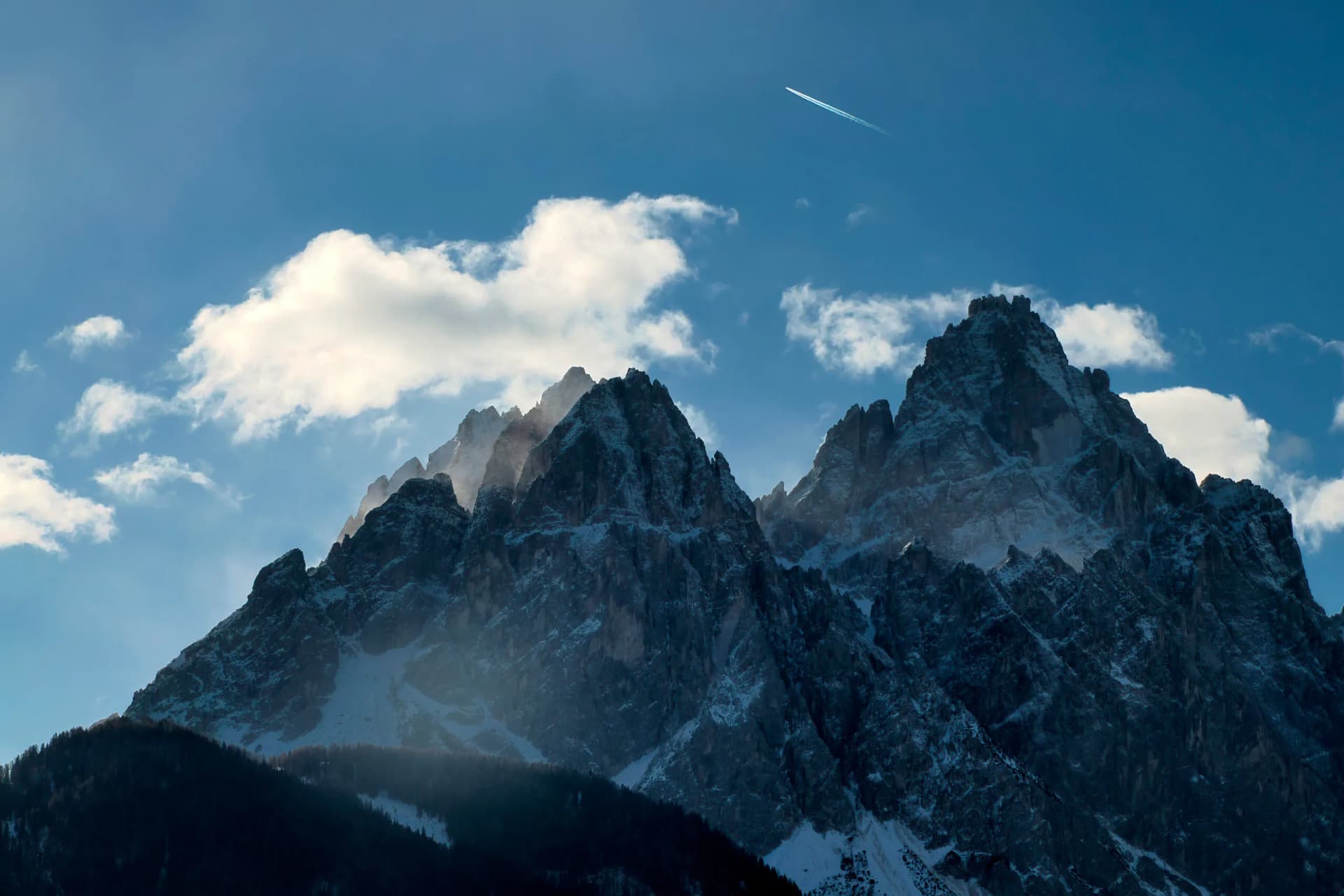 Jagged, snow-dusted Dolomites mountains above dark forest under a bright blue sky with clouds and a jet contrail.
