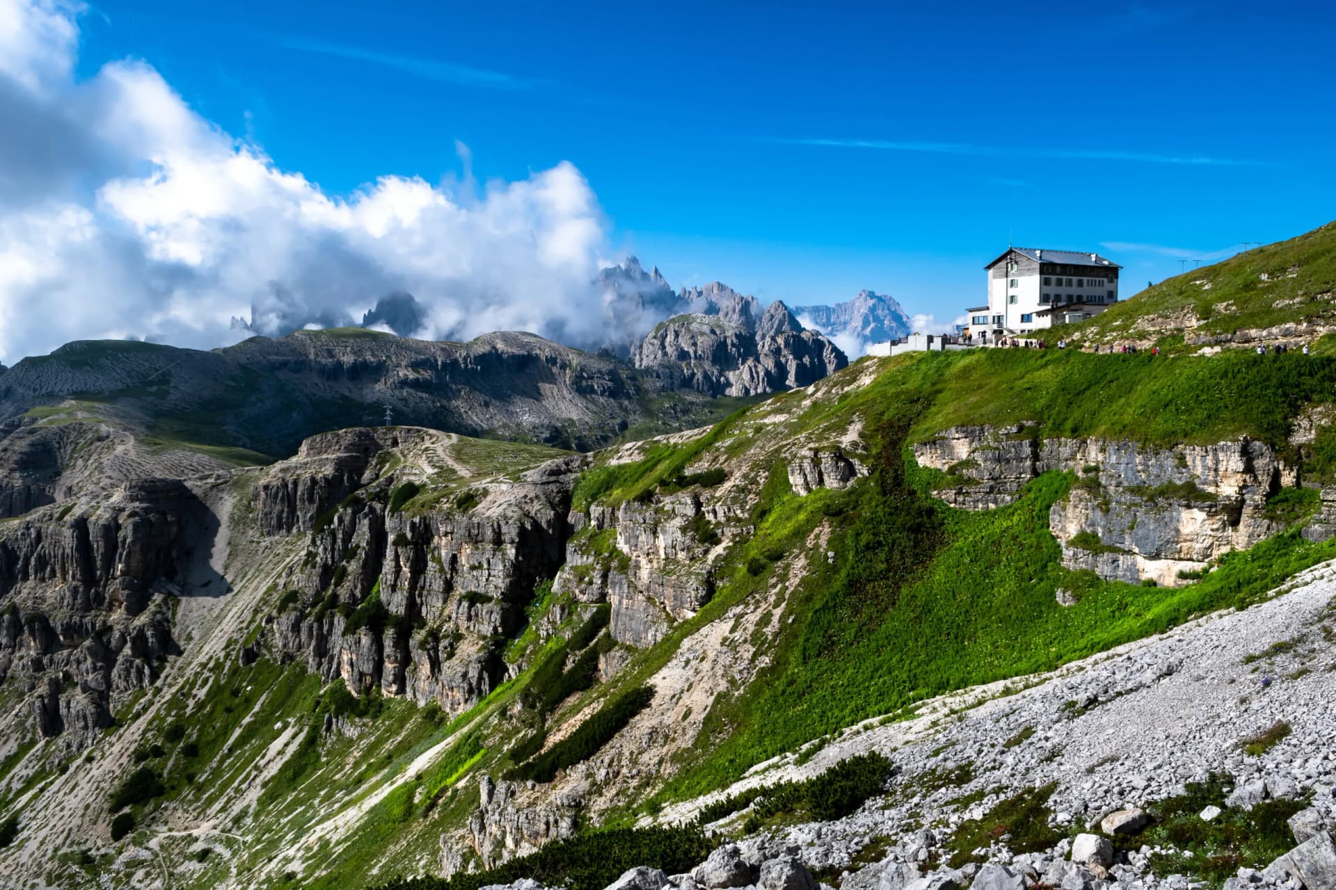 Rifugio Auronzo near Tre Cime with hikers on grassy slope and jagged peaks under blue sky.