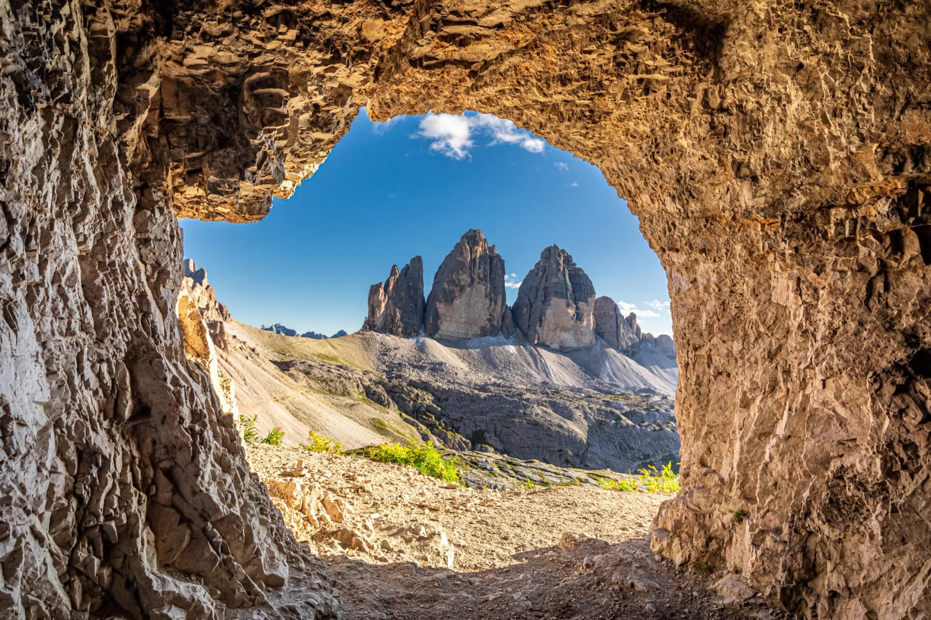 View of Tre Cime di Lavaredo peaks through a rocky cave opening on a sunny day.