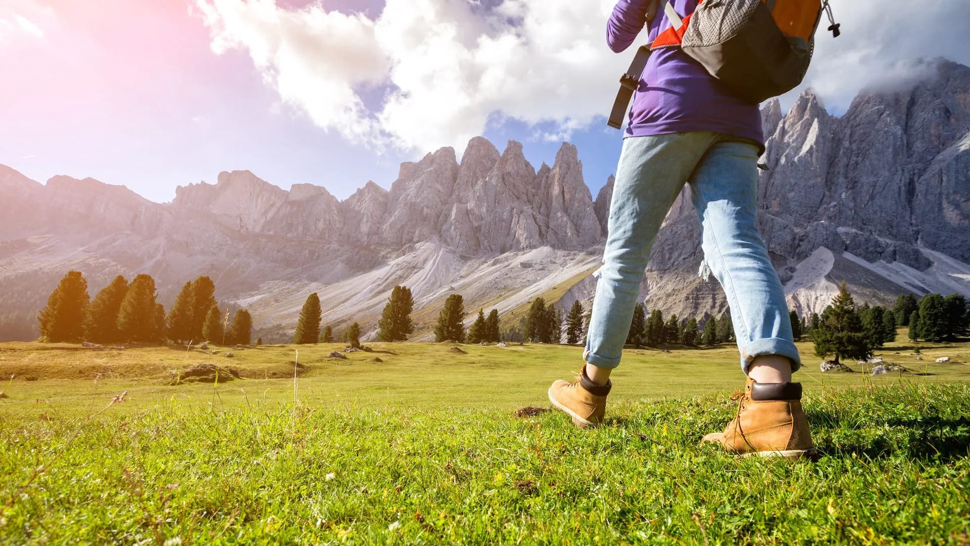 Hiker with backpack walking across grassy meadow toward jagged mountain range