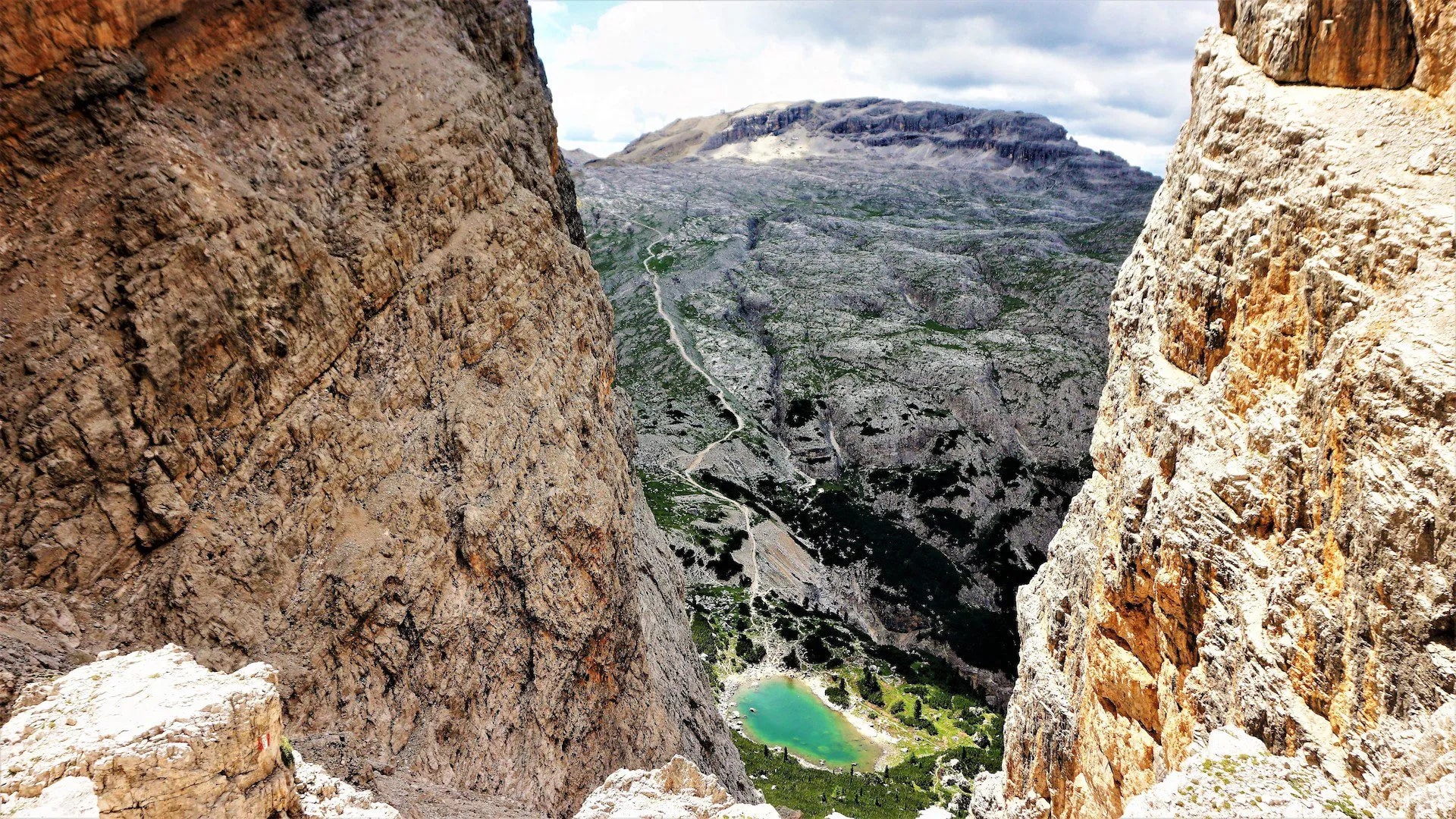 View from Forcella di Lago showing steep rock walls, a winding trail, and a small turquoise alpine lake below.