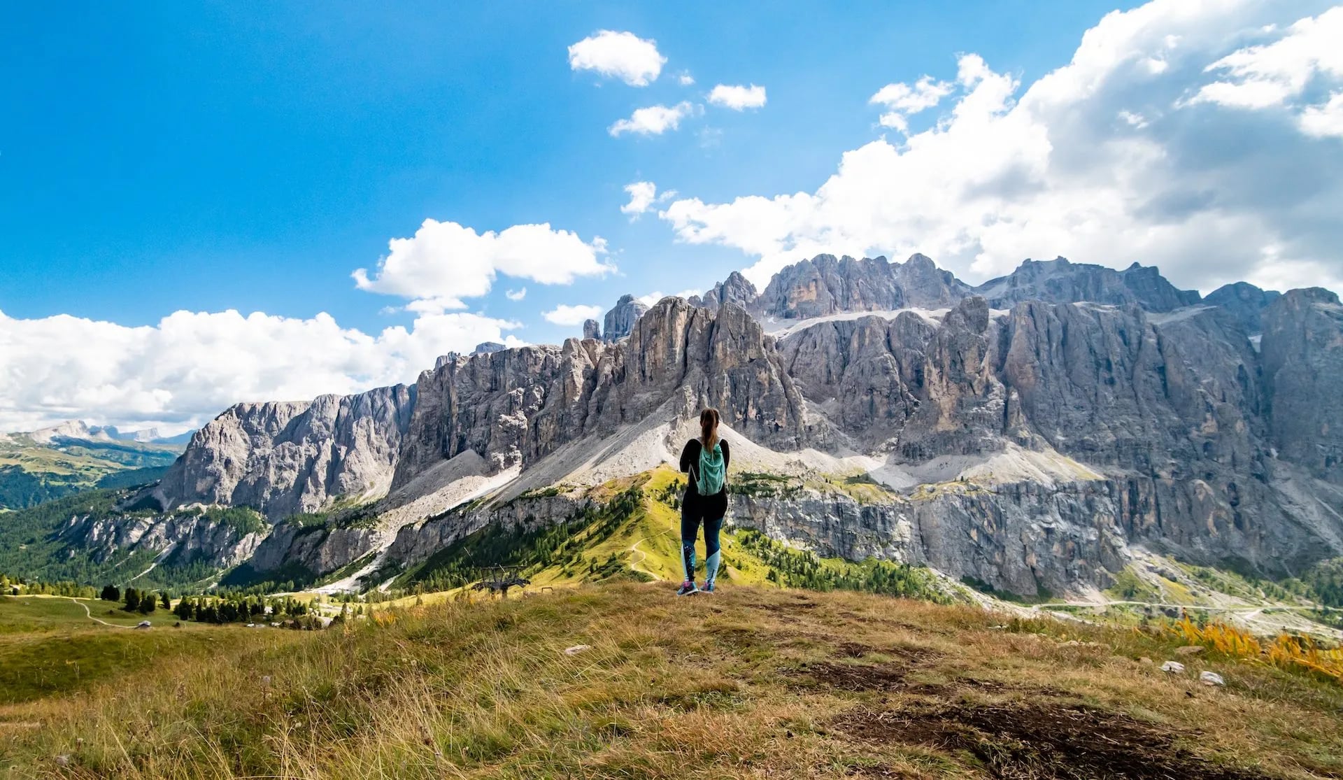 Hiker with backpack standing on grassy overlook facing massive rocky mountains under blue sky.