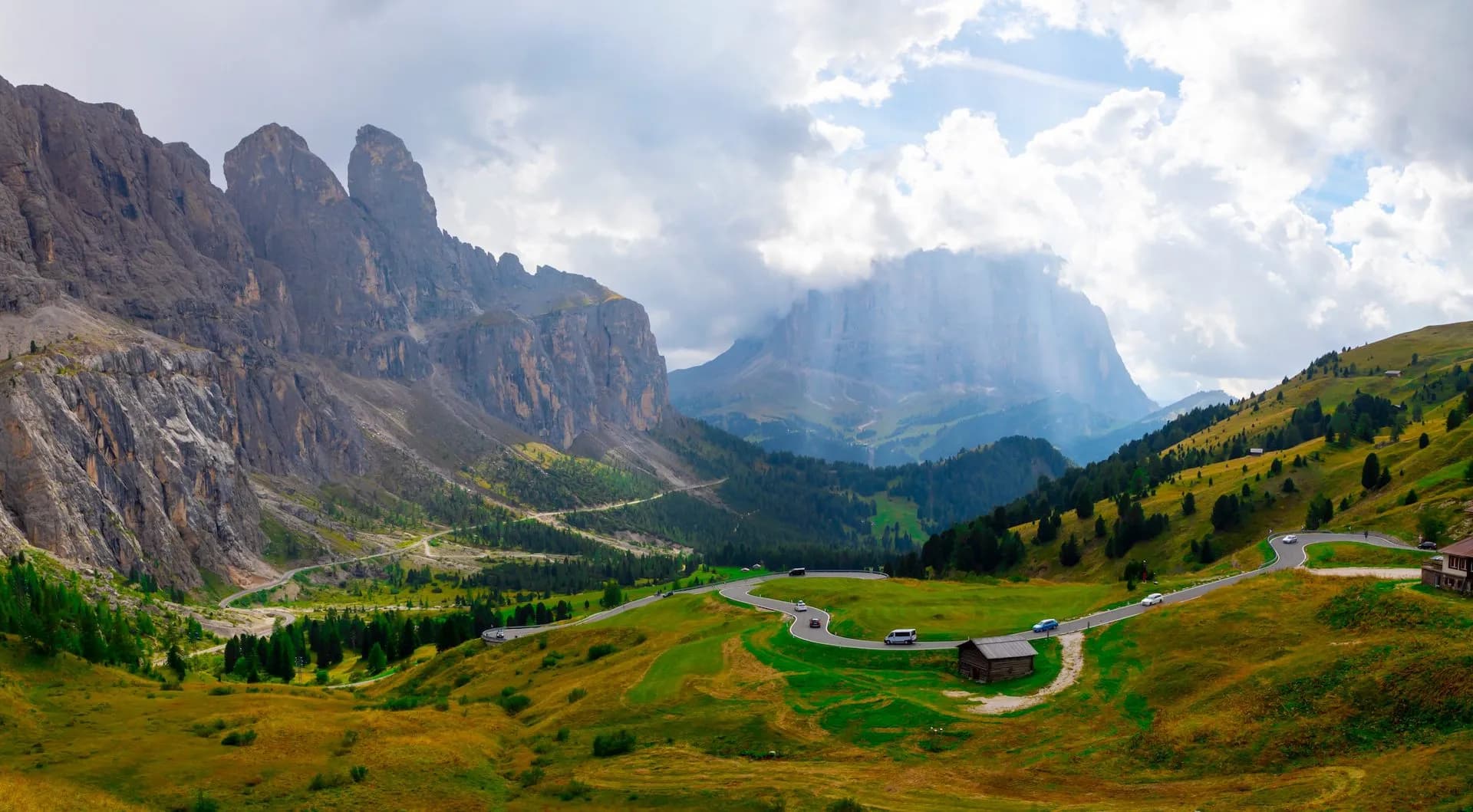 Winding mountain road with cars passing through green and yellow alpine meadows near rocky peaks.