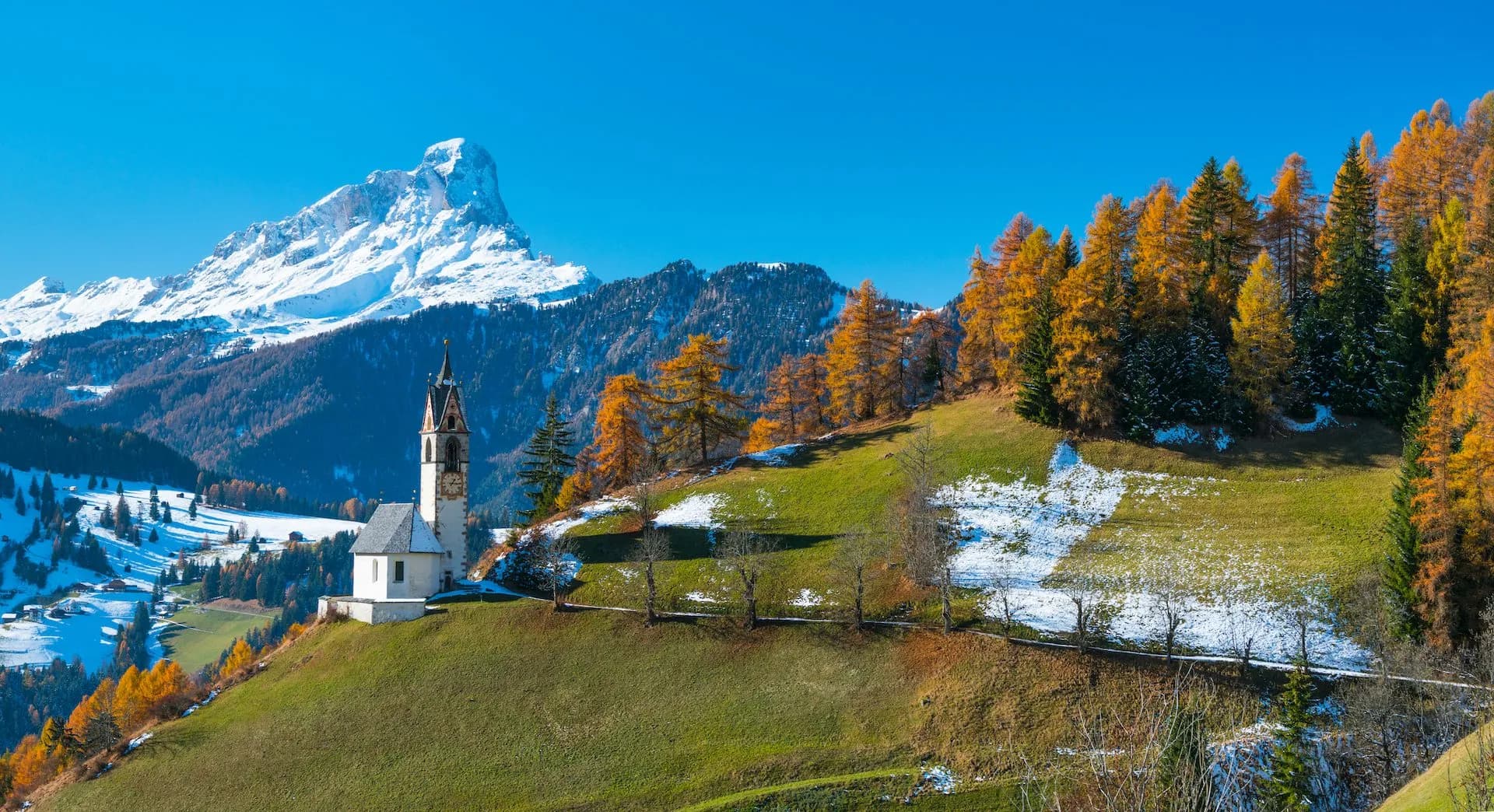 White church on green hillside with autumn trees and snowy mountain backdrop, La Val