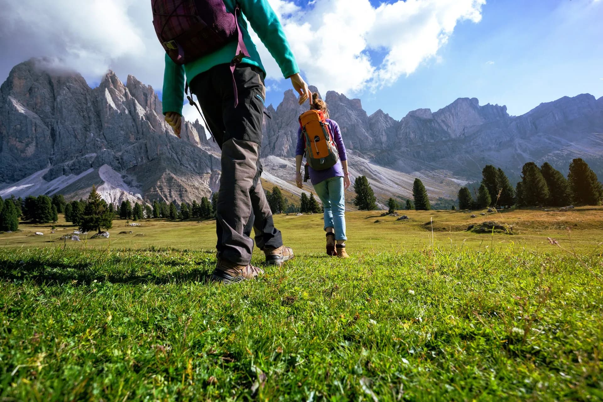 Hikers walking across a grassy meadow toward jagged Dolomite mountains under a blue sky.