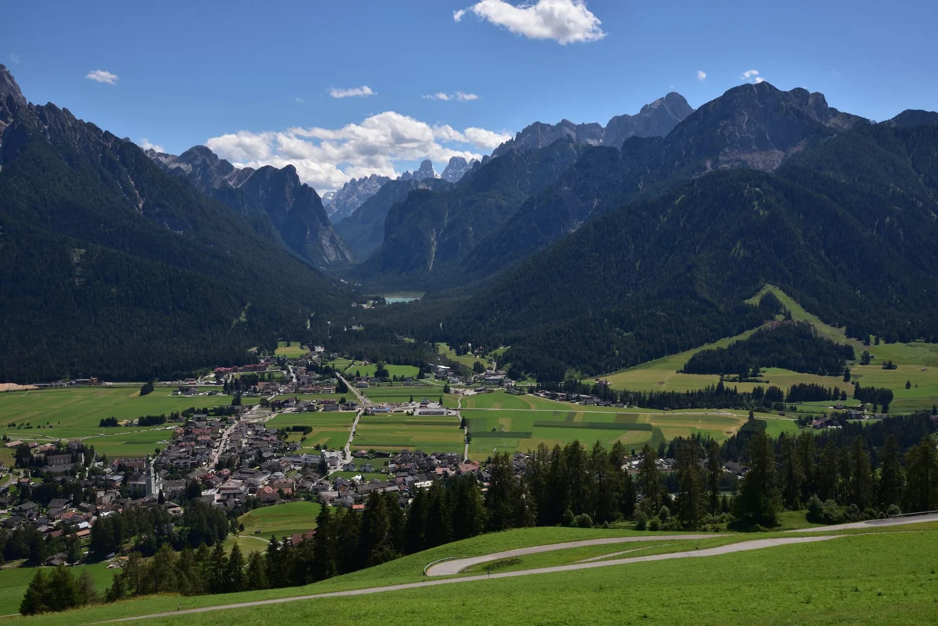 Alpine valley town with green fields and dark forested mountains under blue sky, Val Pusteria and Dobbiaco.