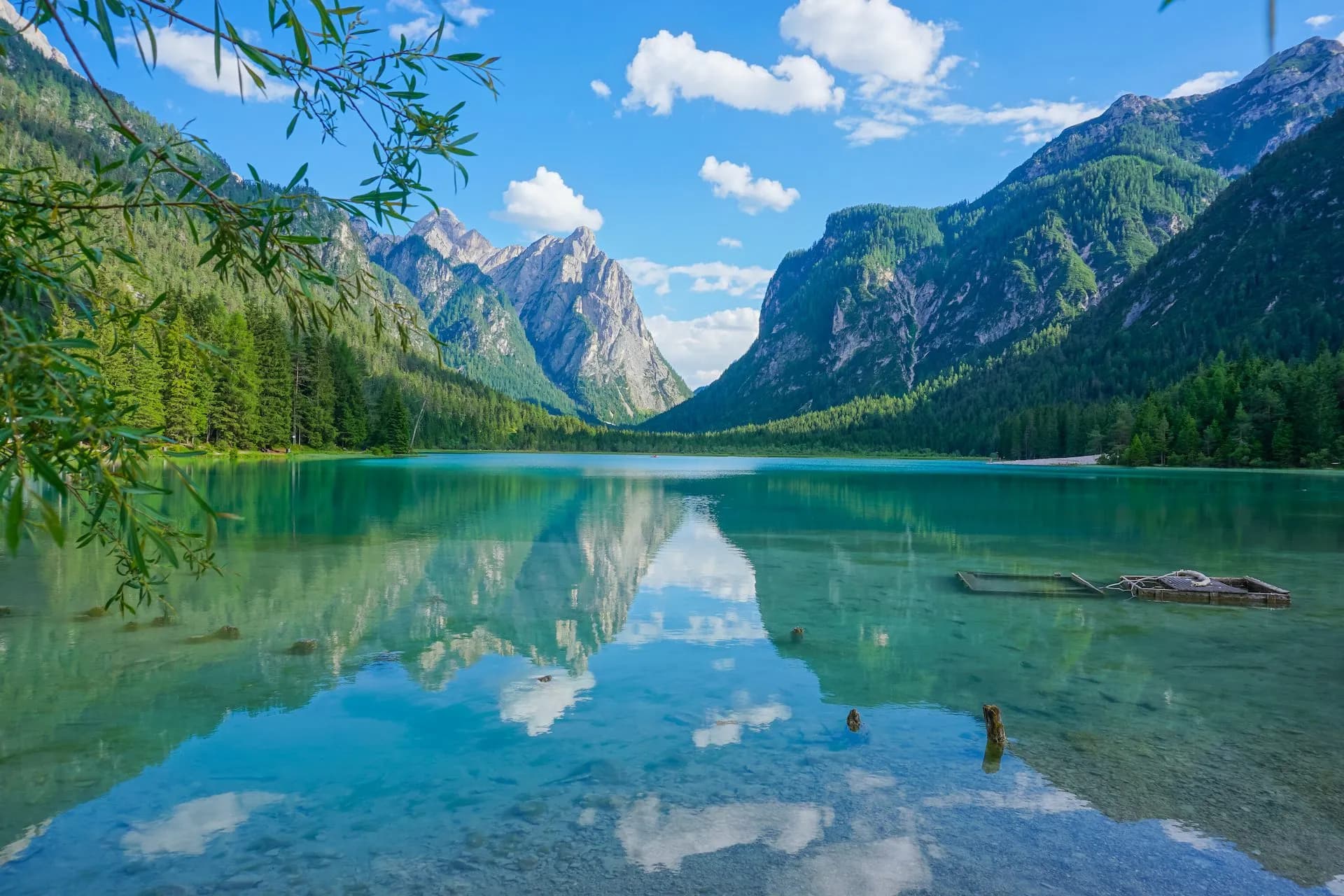 Lake Dobbiaco with turquoise water reflecting steep, forested mountains under a blue sky.
