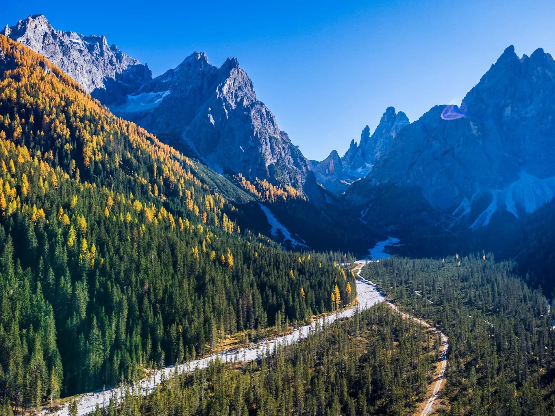 Alpine valley with rugged mountains, autumn trees, and a dry riverbed under clear blue sky.