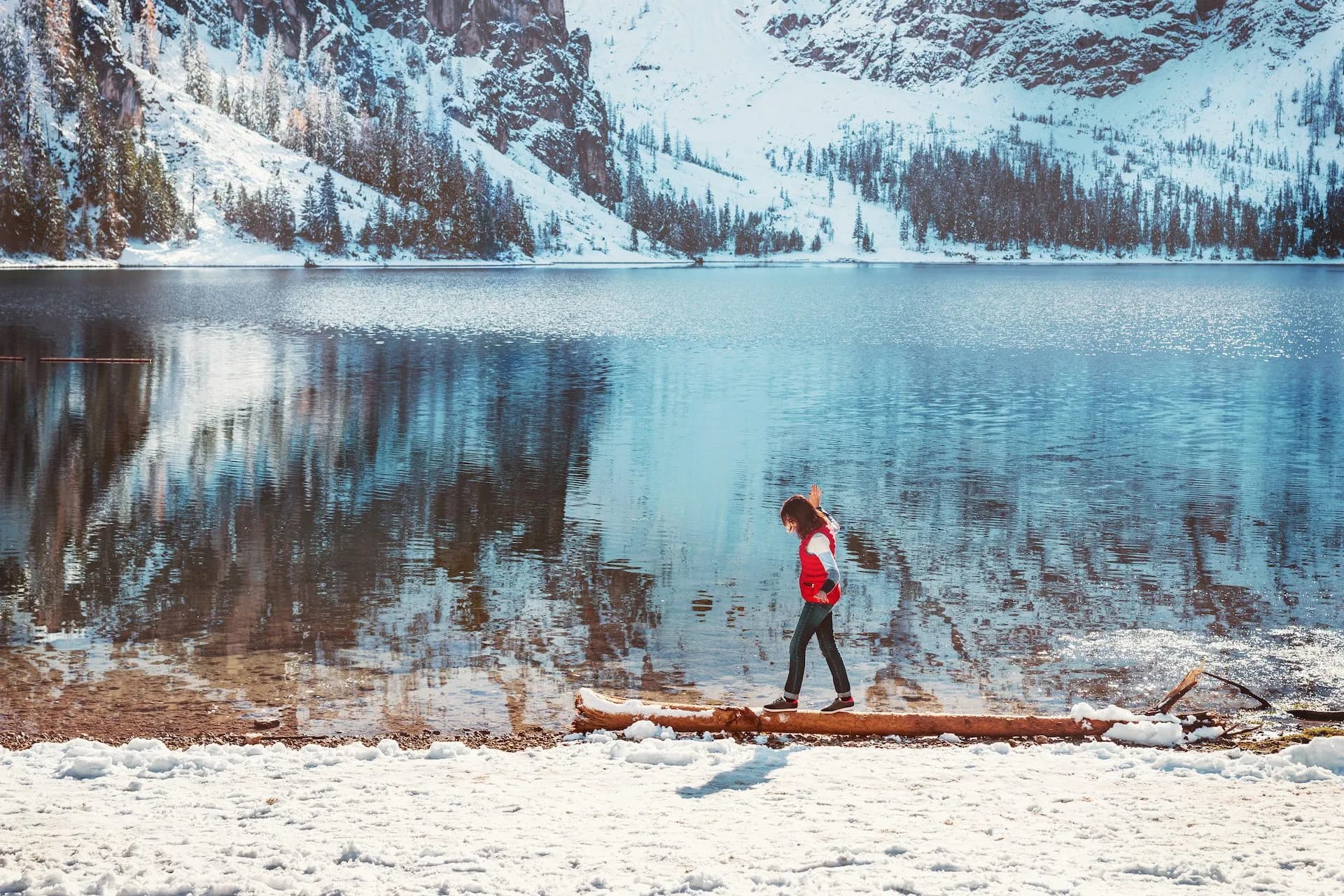 Hiker balances on log by Lago di Braies with snow-covered mountains and blue water