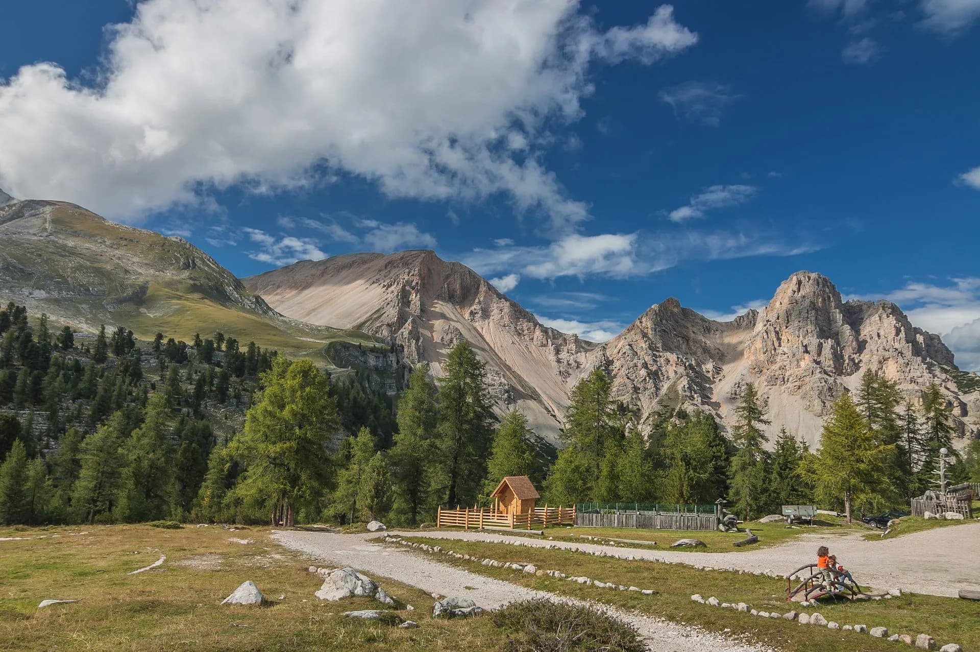 Alpine scenery with rugged mountains, green forest, and a small wooden hut under a blue, cloudy sky.