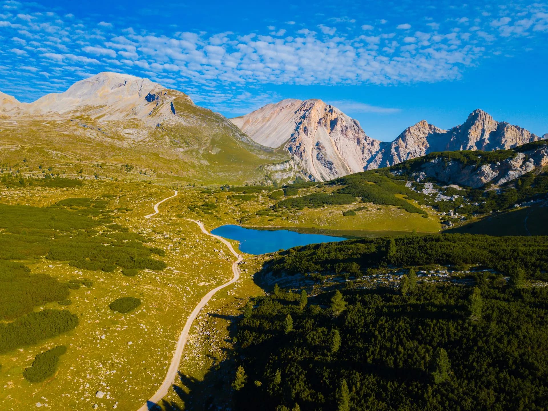 Alpine lake with bright blue water, winding dirt path, and rugged mountains under blue sky.
