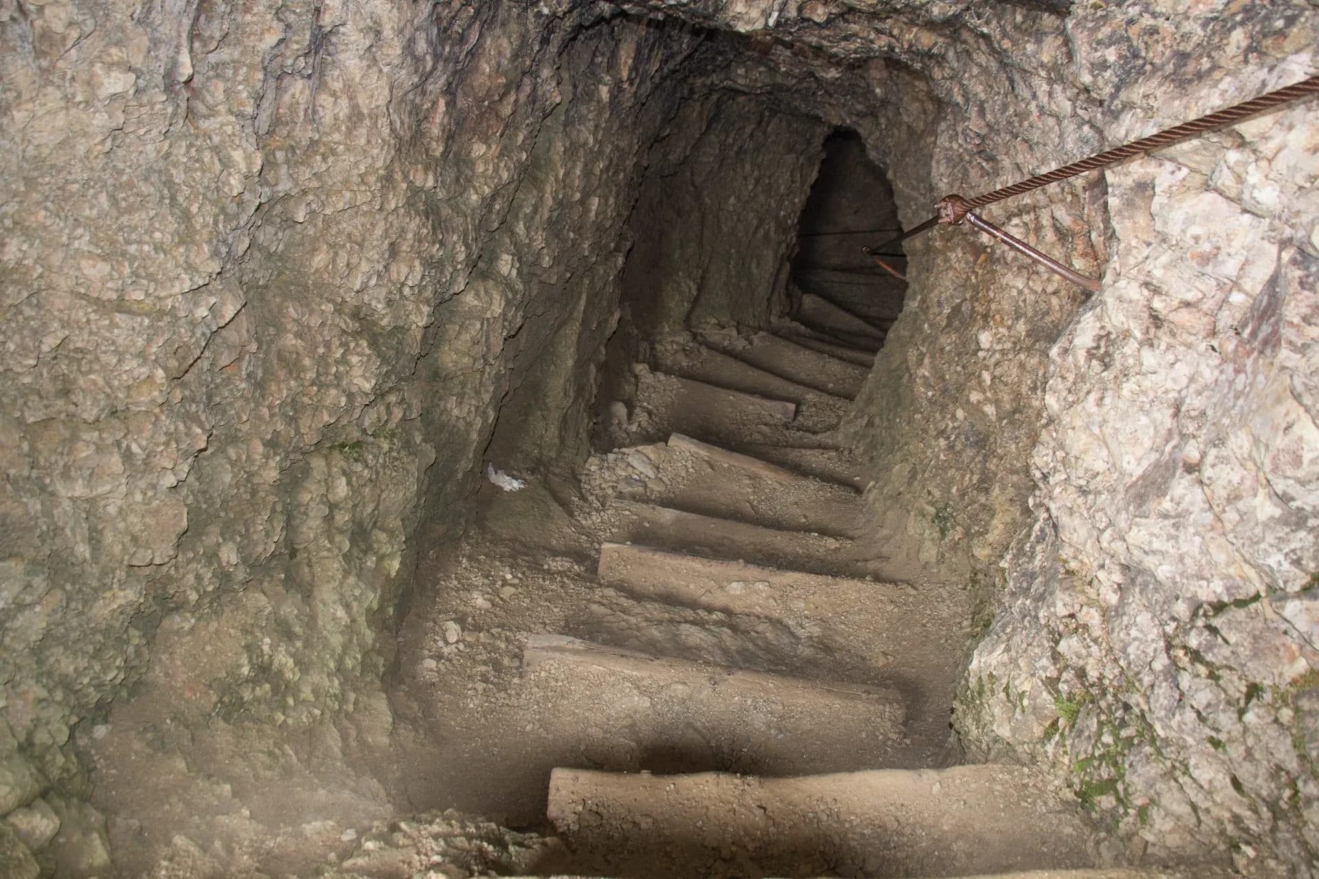 Descending steep, rough-hewn steps inside the Lagazuoi tunnels with a metal cable handrail.