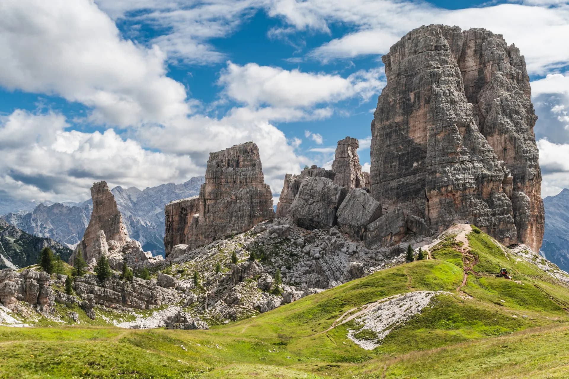 Majestic rock towers of Cinque Torri in the Dolomites with hikers on a grassy trail under a blue sky.
