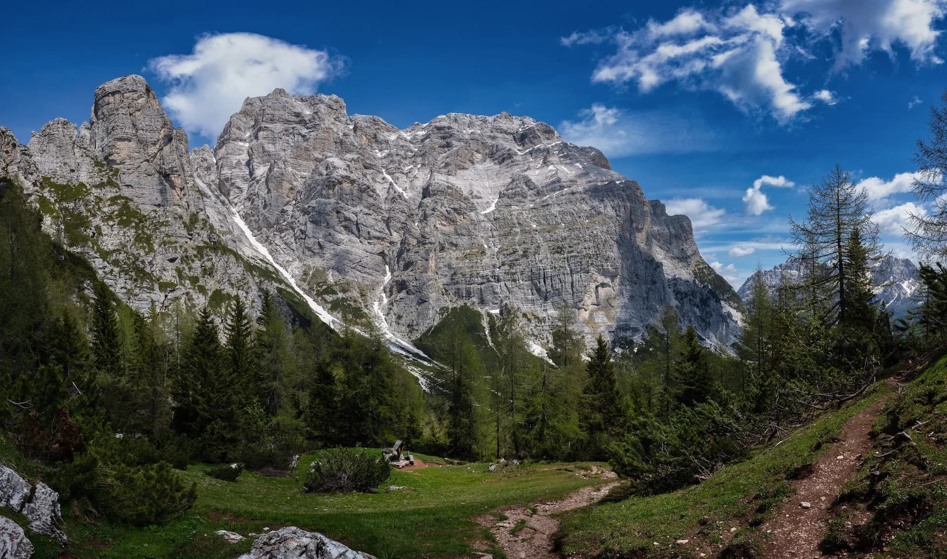 Hiking trail leading toward massive gray mountain peaks with patches of snow under a blue, cloudy sky.