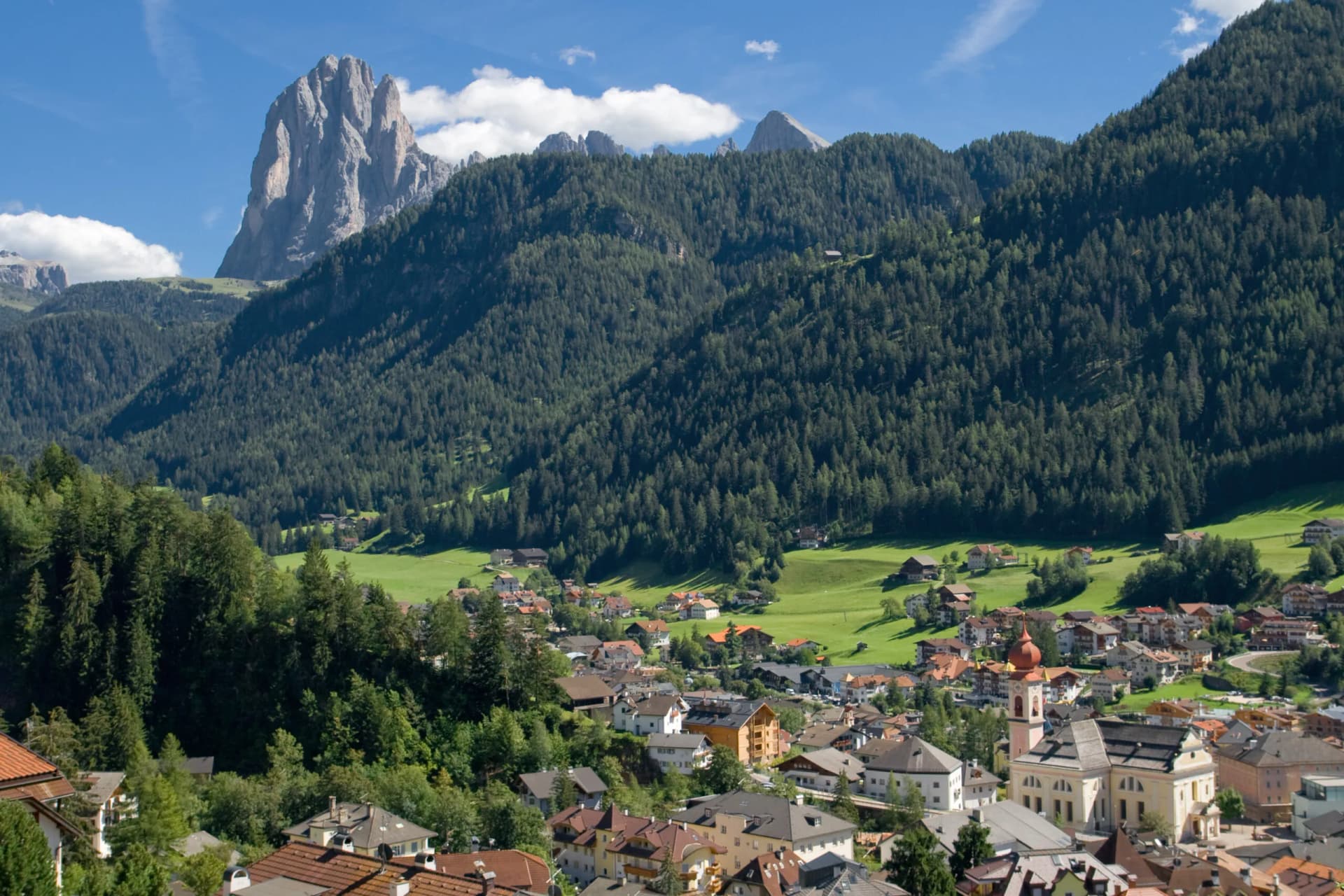 Alpine village nestled in valley with dense forest and Sassolungo mountain peak in background.